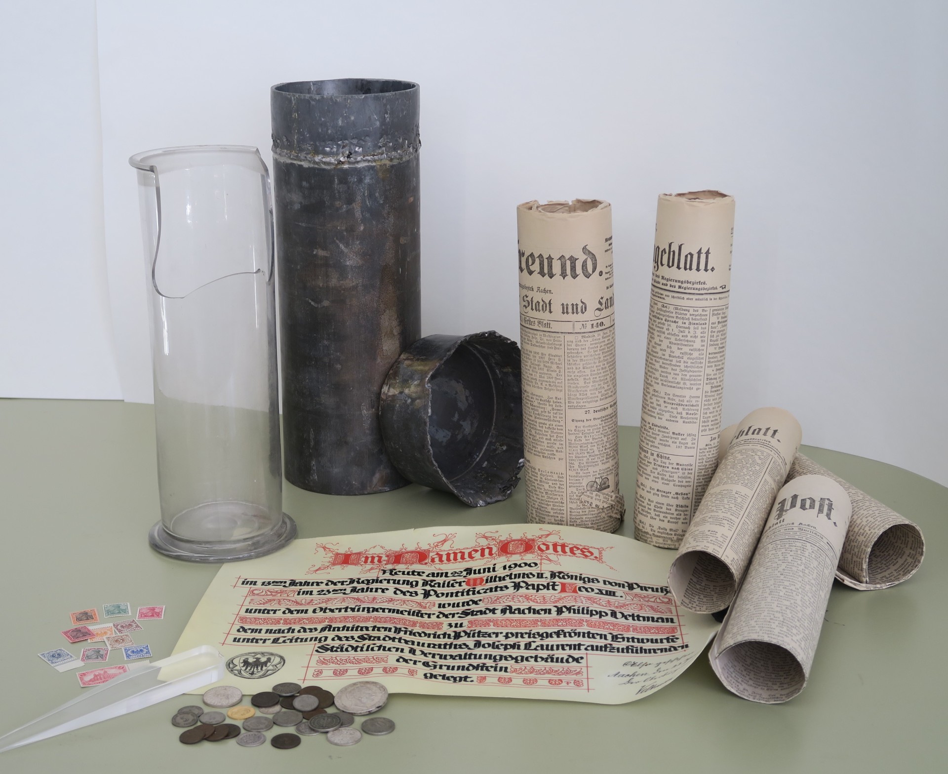 Een glazen cilinder, een loden cilinder met deksel, opgerolde historische kranten, een hostorisch document, munten en postzegels op een tafel.