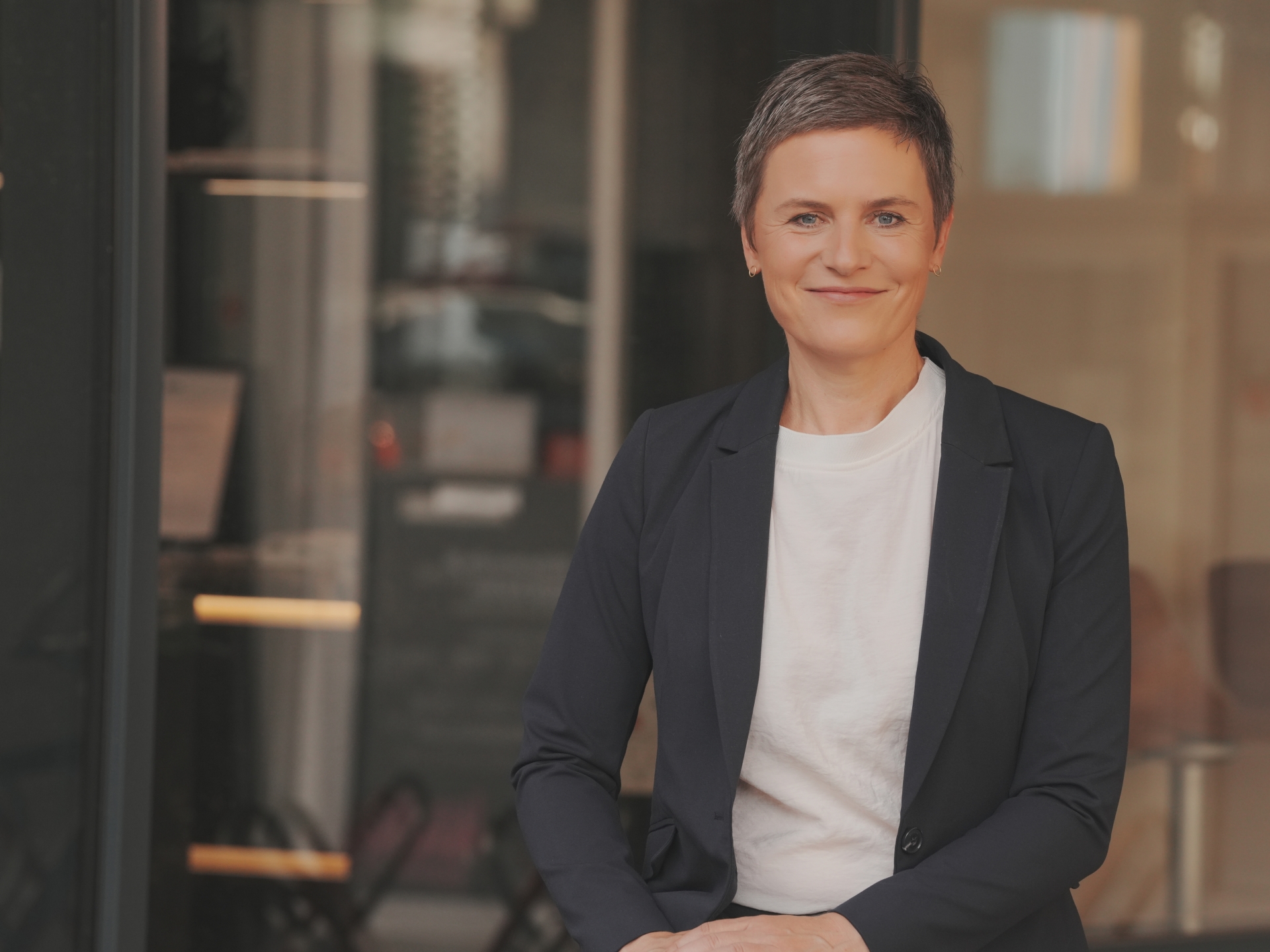 Een vrouw zit voor een groot raam en kijkt in de camera.