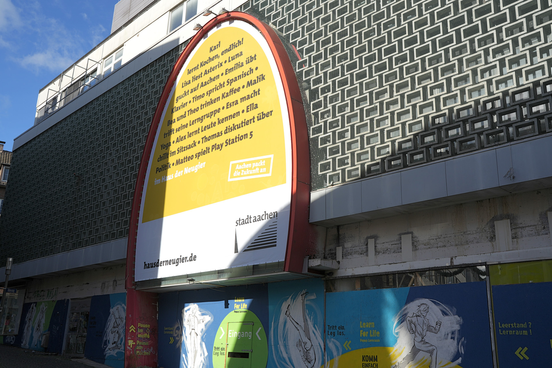 Je ziet een groot ovaal spandoek aan de gevel van een voormalig warenhuis hangen. De banner is een voorbeeld van wat er in de toekomst in het Huis van de Wetengierigheid kan worden gedaan.