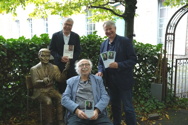 Drie mannen houden de flyer voor het voorprogramma van de Hasenclever Prize vast naast een sculptuur van Walter Hasenclever voor een heg.
