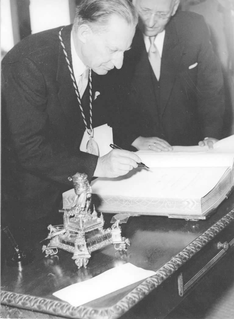 Een zwart-wit foto van de toenmalige Italiaanse president Alcide de Gasperi. Gasperi schrijft met een pen in het gouden boek van de stad Aken.