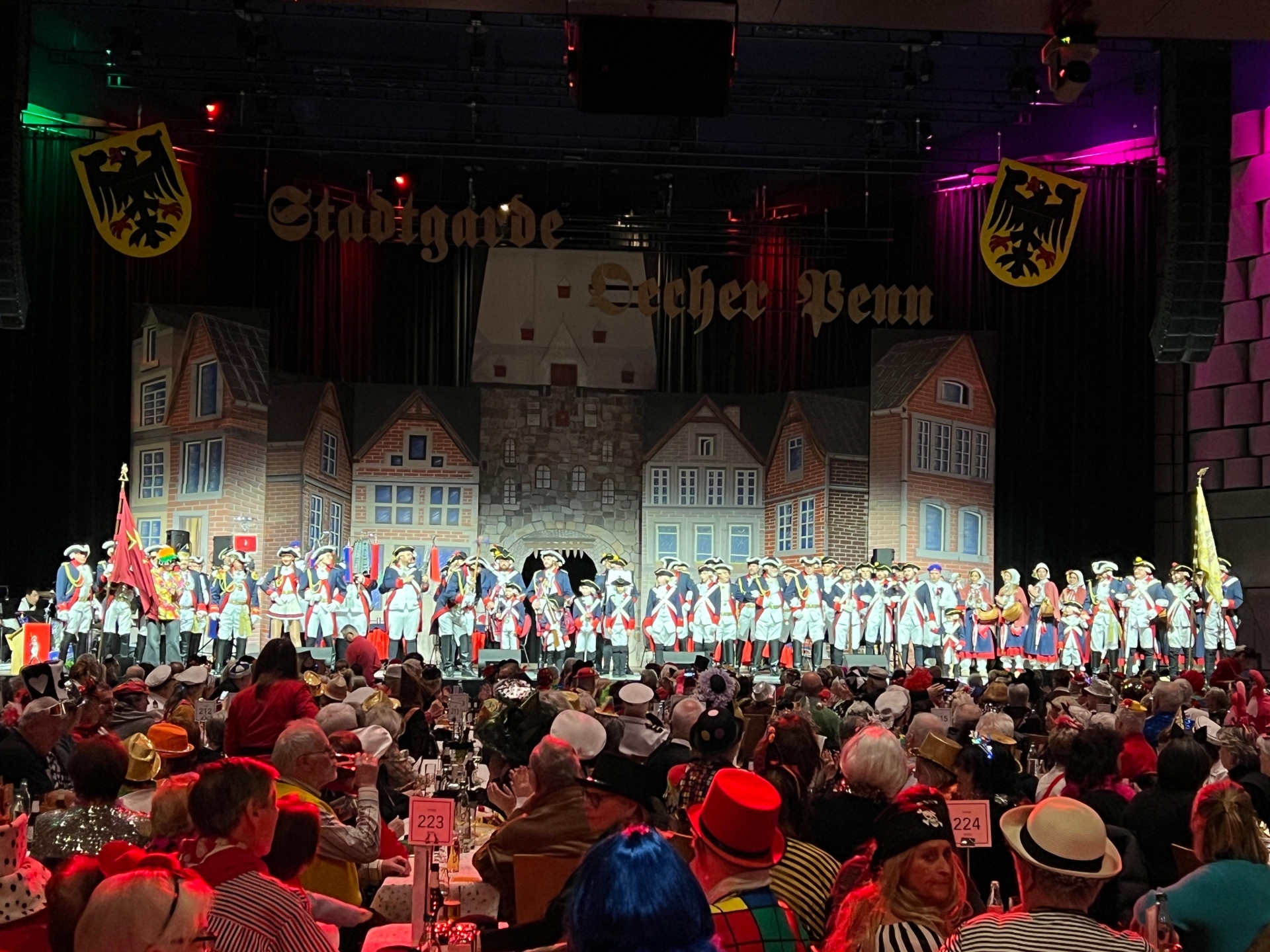 Een volle evenementenhal met mensen in carnavalskleding. Een skyline van de stad op de achtergrond op het podium, met geüniformeerde wachters van de stadswacht van Öcher Penn ervoor.