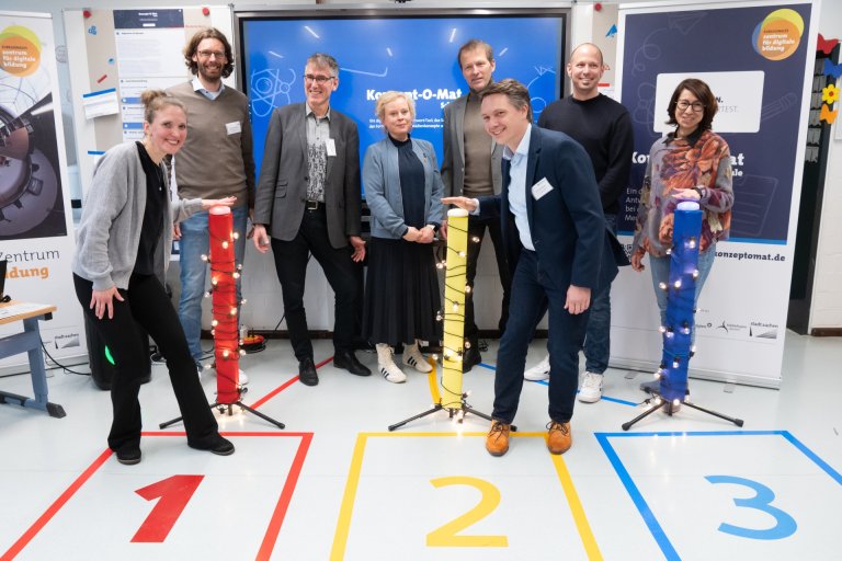 Een groep mensen staat voor drie kleurrijke pilaren met zoemers die de spelshow "1,2 of 3" nabootsen.