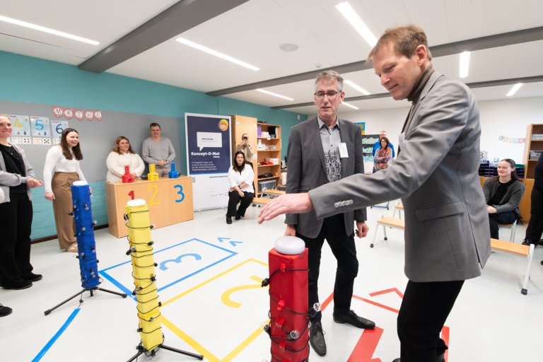 Twee mannen in pak staan voor een rode pilaar met een zoemer. Degene rechts drukt op de zoemer.