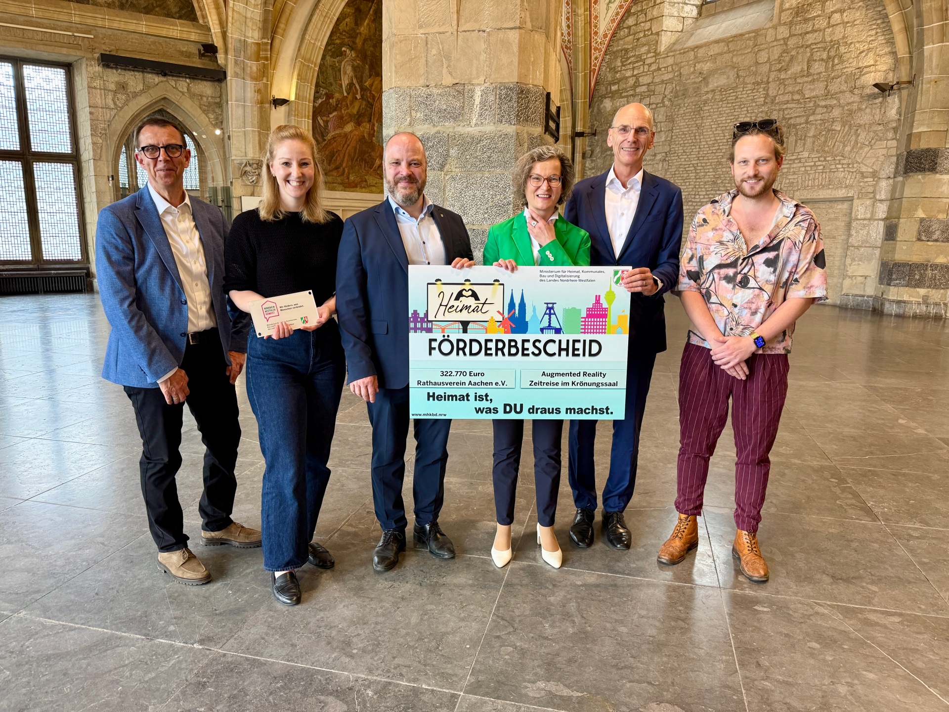 Zes mensen staan in de kroningszaal in Aken en houden een groot subsidiebesluit in hun handen.