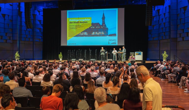 Op de achtergrond: vier mannen staan op een podium en praten. Op de voorgrond: veel rijen stoelen met mensen.