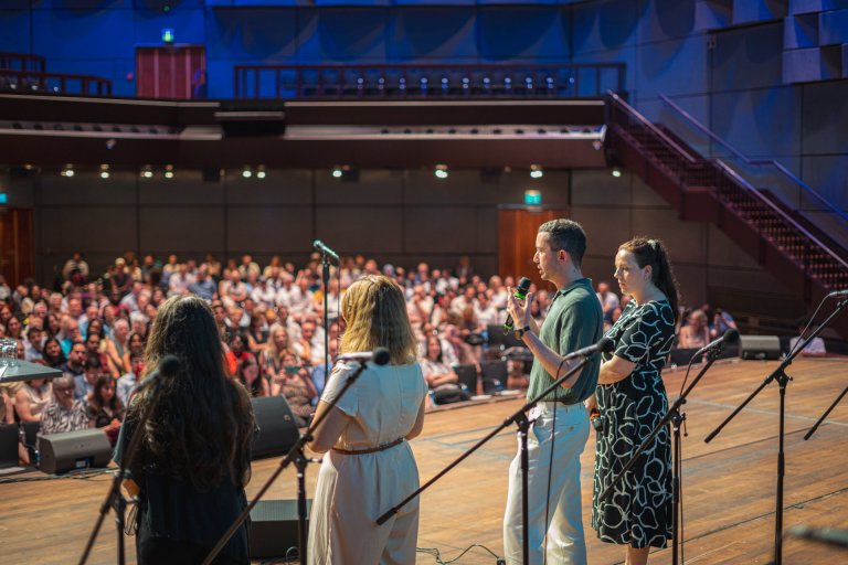 Vier mensen staan op een podium met hun rug naar de camera en praten in microfoons. Op de achtergrond zie je een zaal vol mensen
