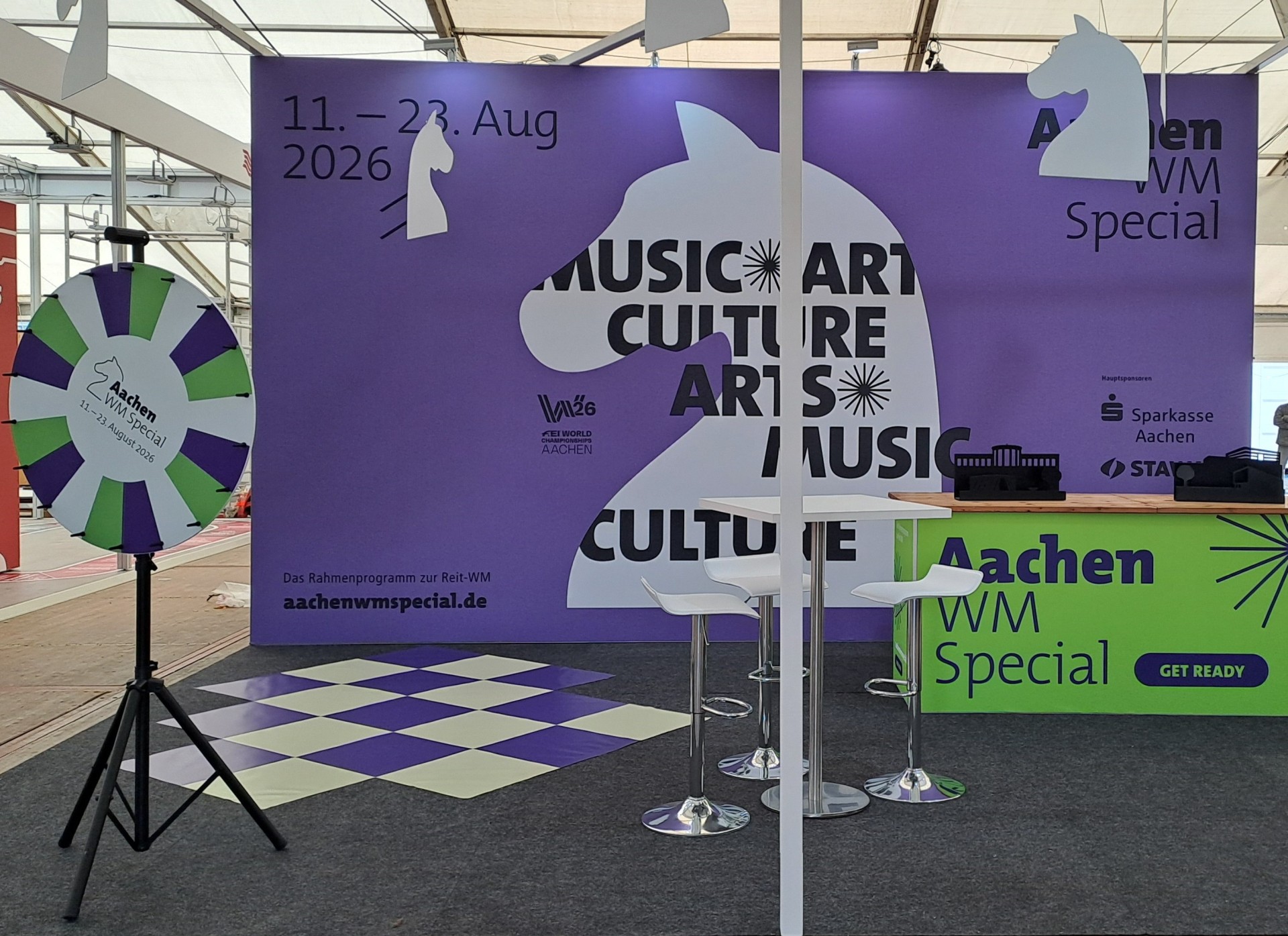 Een beursstand in de kleuren paars, groen en wit, met een rad van fortuin, een tafel met stoelen en witte paarden die aan het plafond hangen.