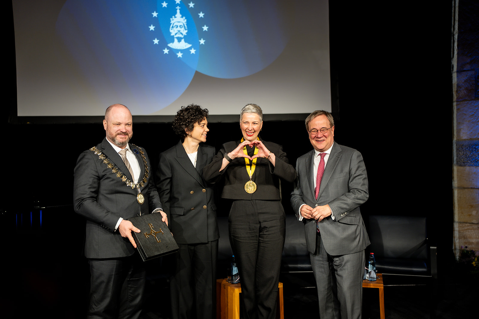 Vier mensen staan op het podium. Een blonde vrouw met de medaille van de Karelsprijs om haar nek toont een hart.