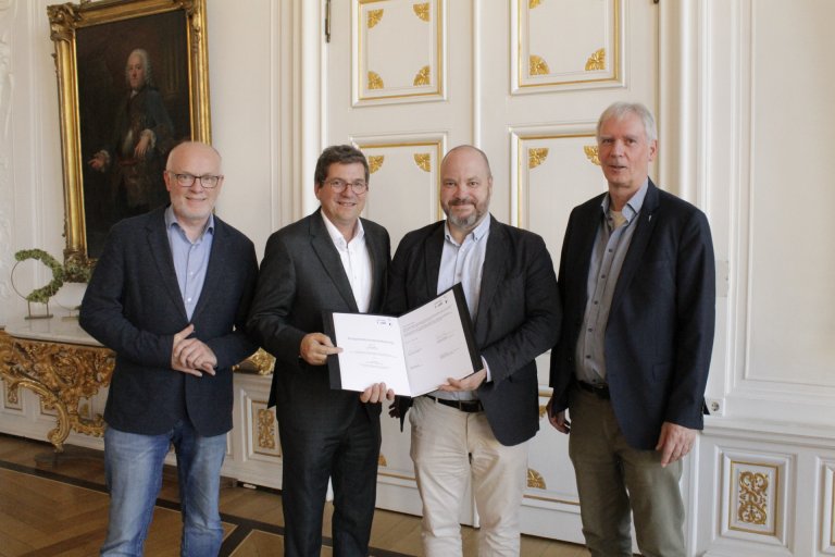 Vier mannen staan voor een witte muur en houden een document in hun handen.