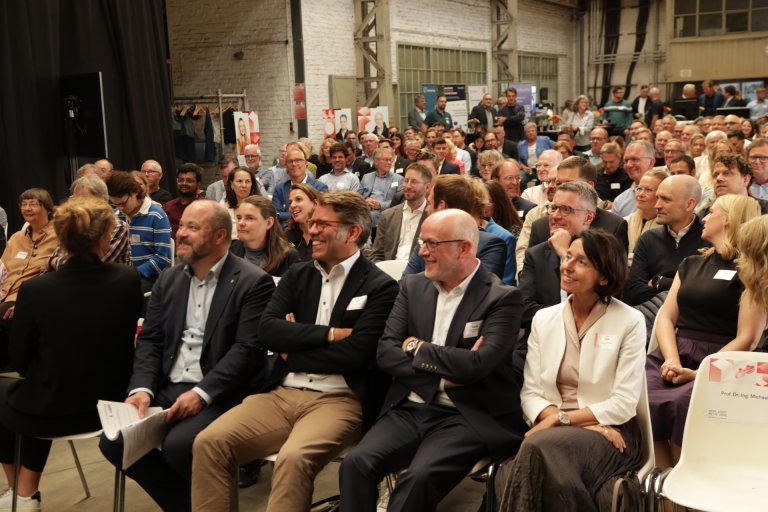 Veel mensen zitten op rijen stoelen in een grote voormalige industriële hal.