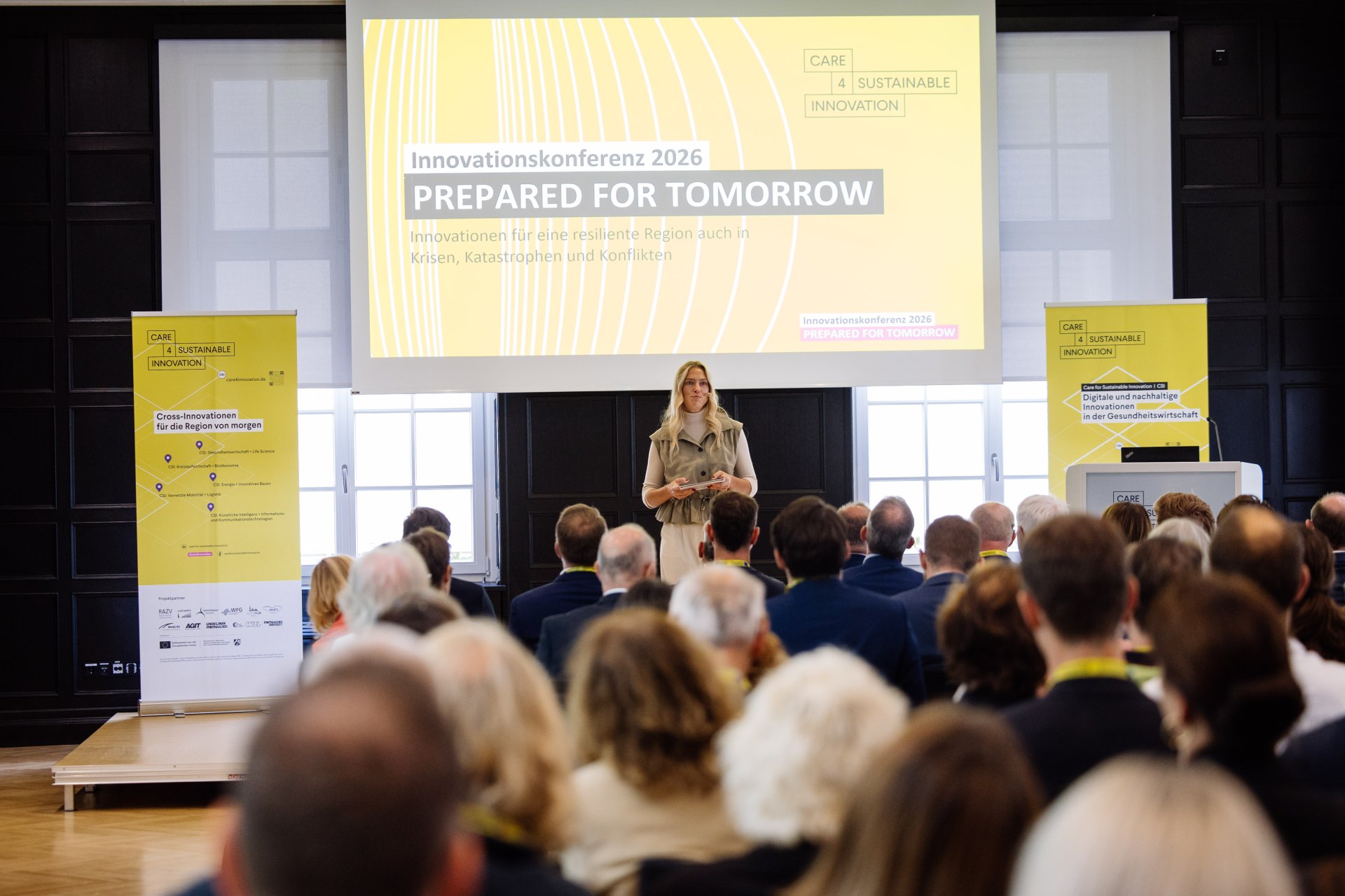 Er staat een vrouw op het podium voor het publiek. Op de achtergrond is een presentatie te zien met de titel "Prepared for Tomorrow"