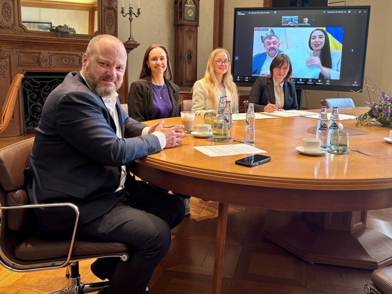 Aan een tafel zit de burgemeester met drie vrouwen en een document. Op de achtergrond staat een groot scherm waarop een videoconferentie te zien is, met een man en een vrouw in beeld.