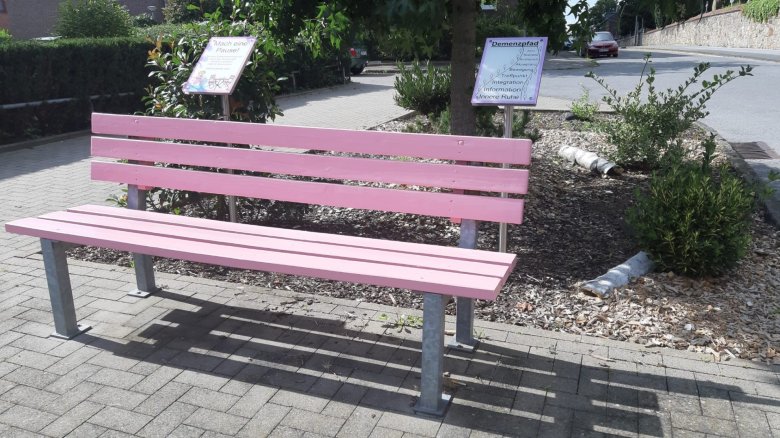 Eine rosa Bank und im Hintergrund zwei Infotafeln mit den Titeln "Demenzpfad" und "Mach eine Pause!"