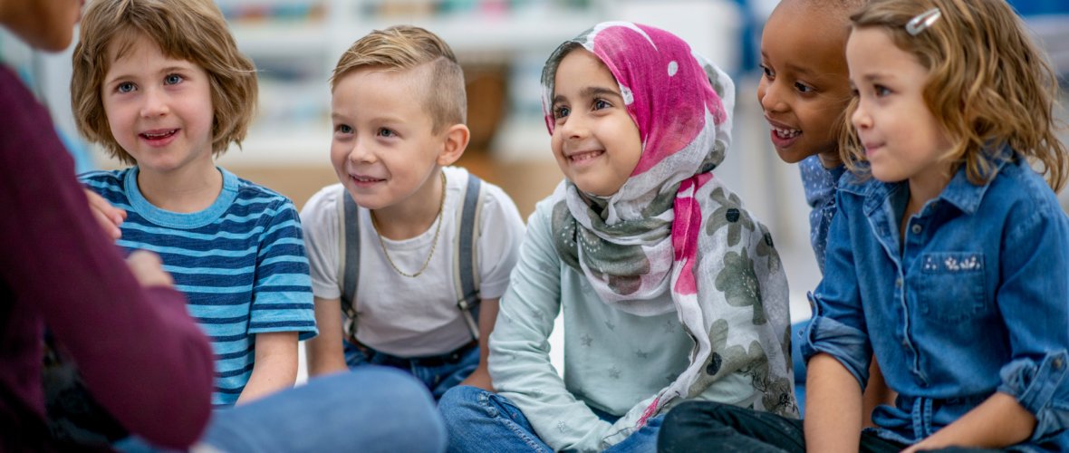 Eine Gruppe Kinder sitzt auf dem Teppich der Klasse und hört einer erwachsenen Person zu