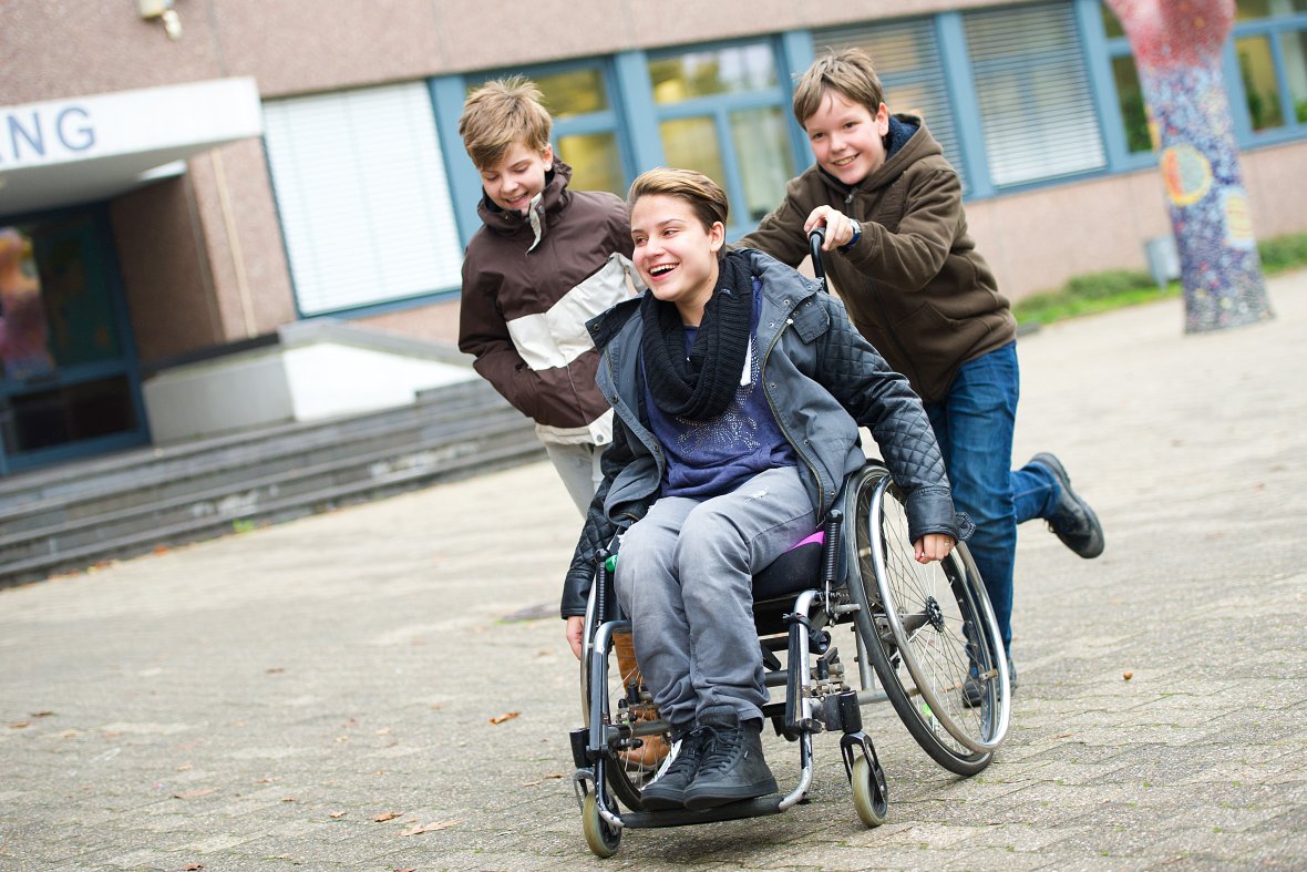Drei Kinder, einer von denen sitzt im Rollstuhl; alle drei lachen