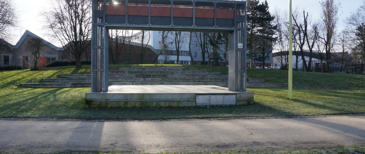 Zu sehen ist die Kennedybühne auf der grünen Wiese des Kennedyparks im Sommer. Die Kennedybühne hat auf diesem Bild ein Dach.