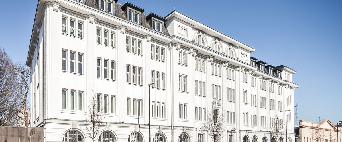 Zu sehen ist die weiße Gebäudefront der Nadelfabrik, davor der Reichsweg, vor blauem Himmel.