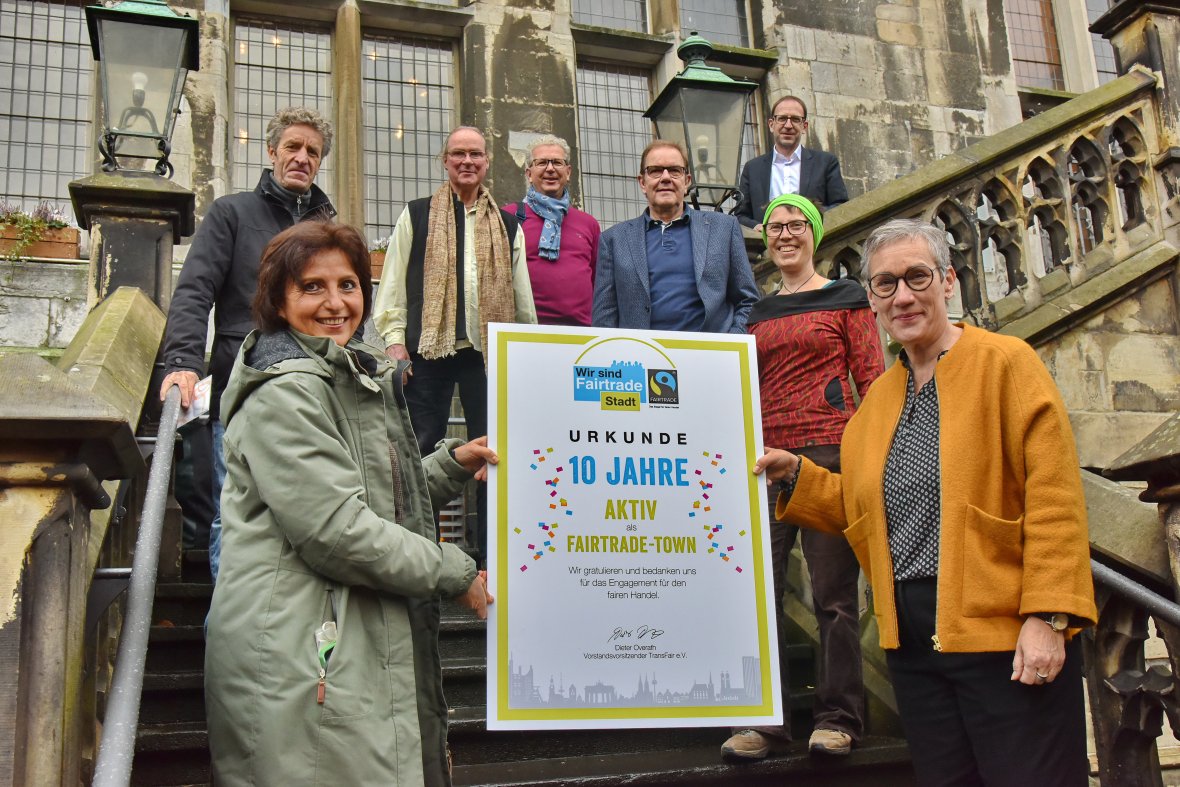 10 Jahre Fairtrade-Town Sibylle Keupen und weitere Beteiligte stehen auf der Rathaustreppe und halten eine Urkunde mit der Aufschrift: Urkunde - 10 Jahre aktiv als Fairtrade-Town" in den Händen.