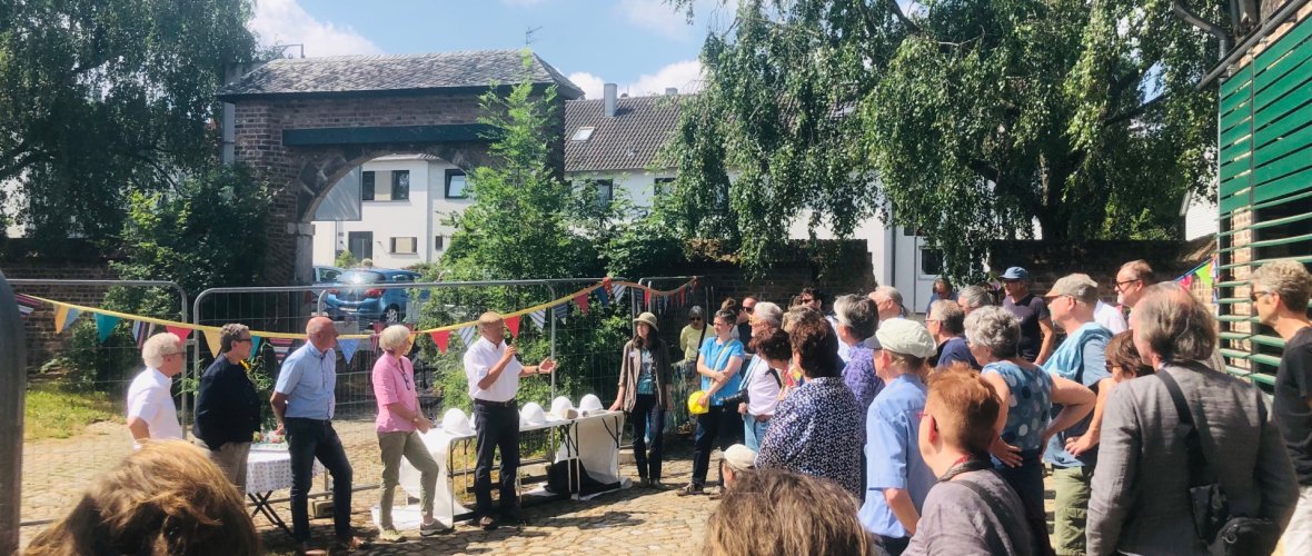 Viele Menschen stehen im Halbkreis gegenüber sechs weiteren Menschen, die vor Tischen mit Bauhelmen stehen. Ein Mann spricht in ein Mikrofon. Im Hintergrund ein ein geschmückter Bauzaun und eine alte Toreinfahrt.
