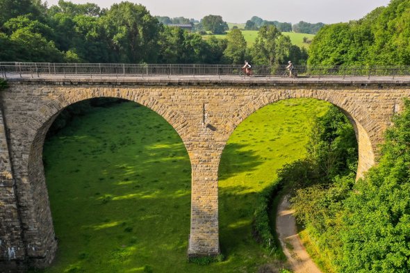 Großes Viadukt, über das mit Fahrrädern gefahren wird, in einer grünen Landschaft mit Wiesen und Bäumen