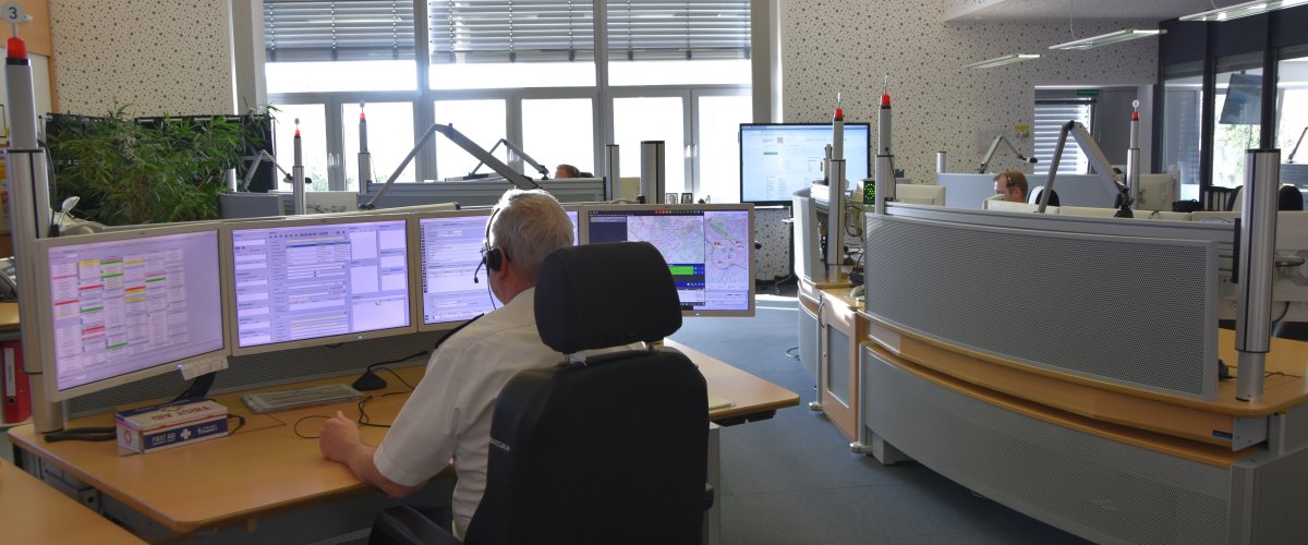 Einblick in die Leitstelle Im täglichen Einsatz sind in der Leitstelle immer mehrere Plätze belegt und weitere jederzeit einsatzbereit. Gerade sitzen vier Mitarbeitende sichtbar an ihren Plätzen und nehmen Notrufe entgegen. Dabei sitzt ein Mitarbeiter mit dem Rücken zur Kamera und hat vor sich diverse Programme auf den Bildschirmen. Unter anderem eine detaillierte Karte von Aachen.