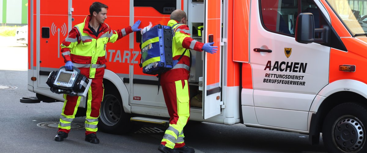 zwei uniformierte Rettungsdienstmitarbeitende sind an der Einsatzstelle eingetroffen, und entnehmen das benötigte medizinische Material zur Versorgung von Patient*innen