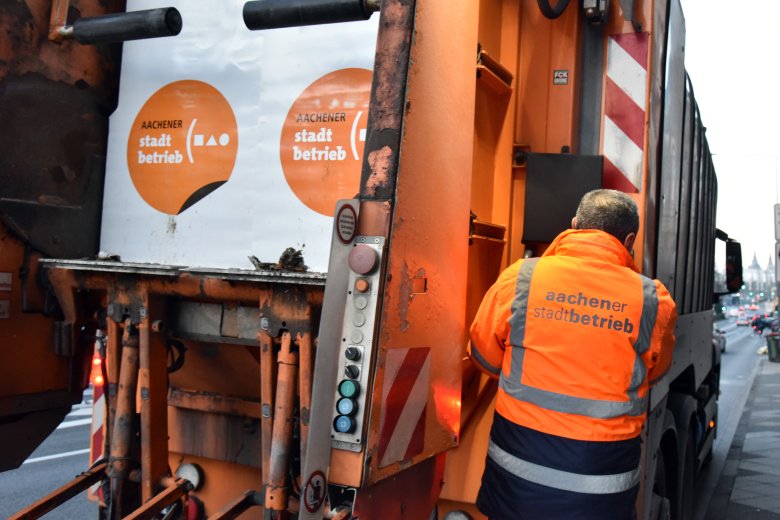 Rückansicht eines Müllfahrzeugs. Ein Müllwerker hält sich rechts am Wagen fest.