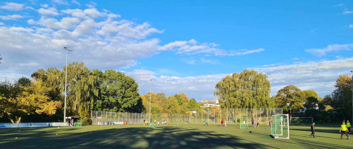 Auf einem Fußballplatz sind mehrere Kleinfelder aufgebaut. Links spielen viele Kinder Fußball. In der Bildmitte eine freie grüne, kurz geschnittene Rasenfläche. Im Hintergrund Fangzäune und Bäume. Blauer Himmel