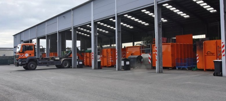 Recyclinghof Eilendorf. Ein Lastwagen mit Haken steht vor einer großen Halle, in der orangene Container stehen.