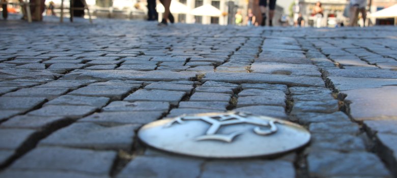 Bodennahe Aufnahme von Kopfsteinpflastersteinen auf dem Markt, mittig eine goldene Plakette mit dem Karlssiegel. Im Hintergrund verschwommen Häuser auf dem Markt.