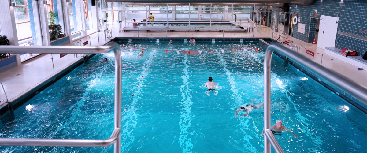 Schwimmhalle Süd Vom Sprungbrett aus sieht man auf das Becken heunter. Im Wasser sind ein paar Schwimmer*innen.