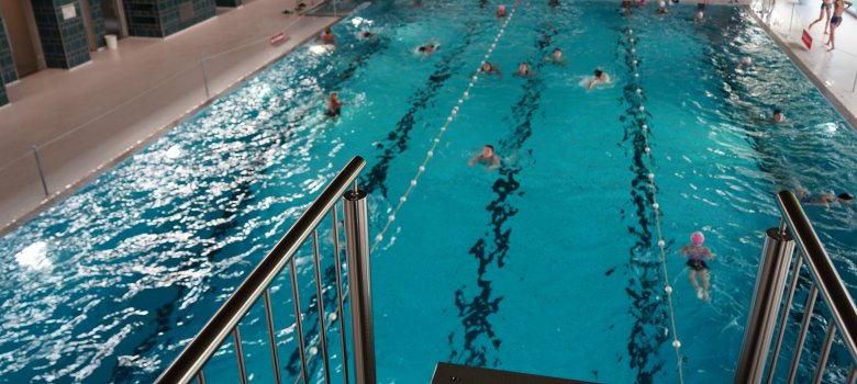 Das Schwimmbecken in der Schwimmhalle Brand vom Springturm aus fotografiert. Im Wasser ziehen mehrere Schwimmer*innen ihre Bahnen.