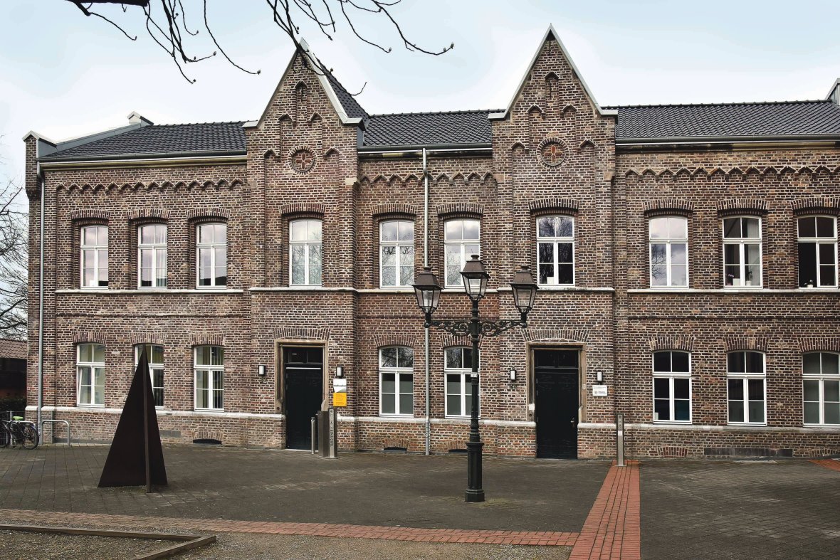 Man sieht den Eingangsbereich des Bezirksamtes. Das Gebäude aus Ziegelsteinen hat zwei spitze Dachgauben. Im Hof stehen außerdem eine schwarze Laterne und ein Fahrrad.