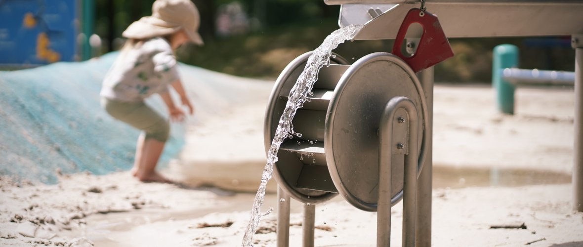 Man sieht ein Wasser-Spielgerät in einem Sandkasten. Das Wasser treibt die Mühle an. Im Hintergrund bückt sich ein Mädchen zu einer Pfütze herunter.