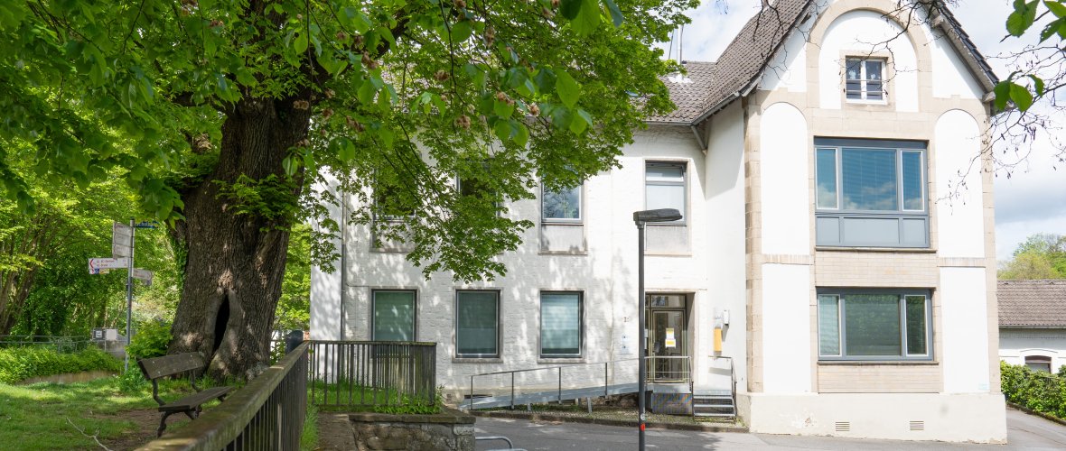 Man sieht das Bezirksamt, ein weißes Gebäude mit einem Spitzdach, und den Parkplatz. Neben dem Amt stehen ein großer grüner Laubbaum und eine Bank. Die Sonne scheint und es sind ein paar Wolken am Himmel.