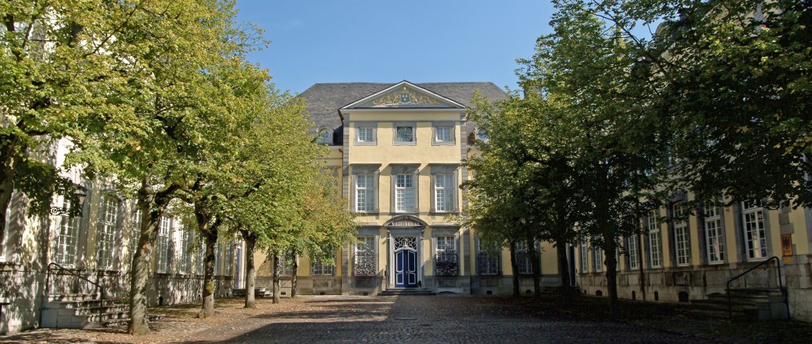 Eine Allee von Bäumen führt zum Eingang der ehemaligen Reichsabtei. Der Himmel ist blau.