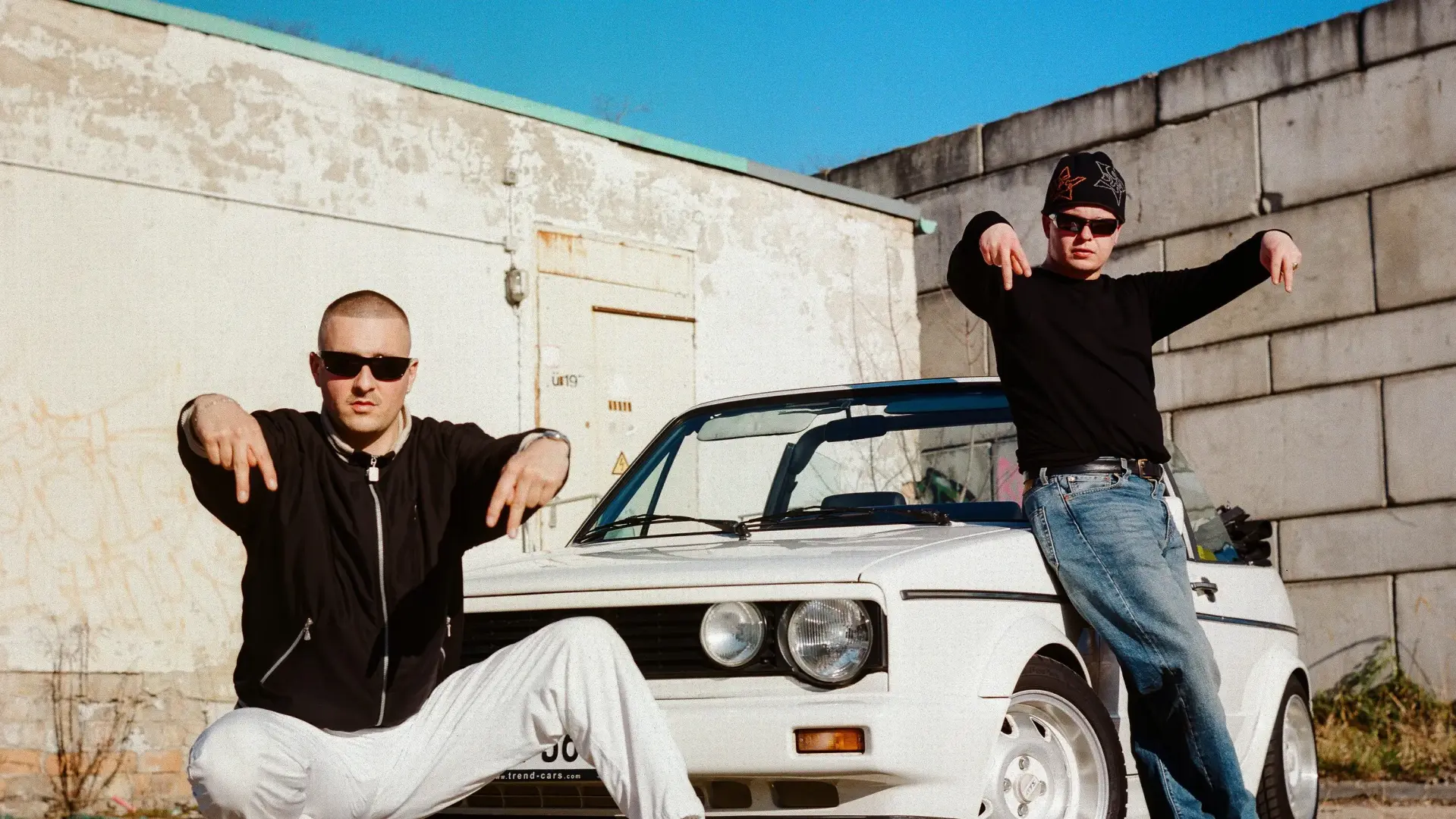 Das Hip-Hop Duo posiert vor einem Auto. Sie tragen Sonnenbrillen und zeigen Symbole mit ihren Händen.