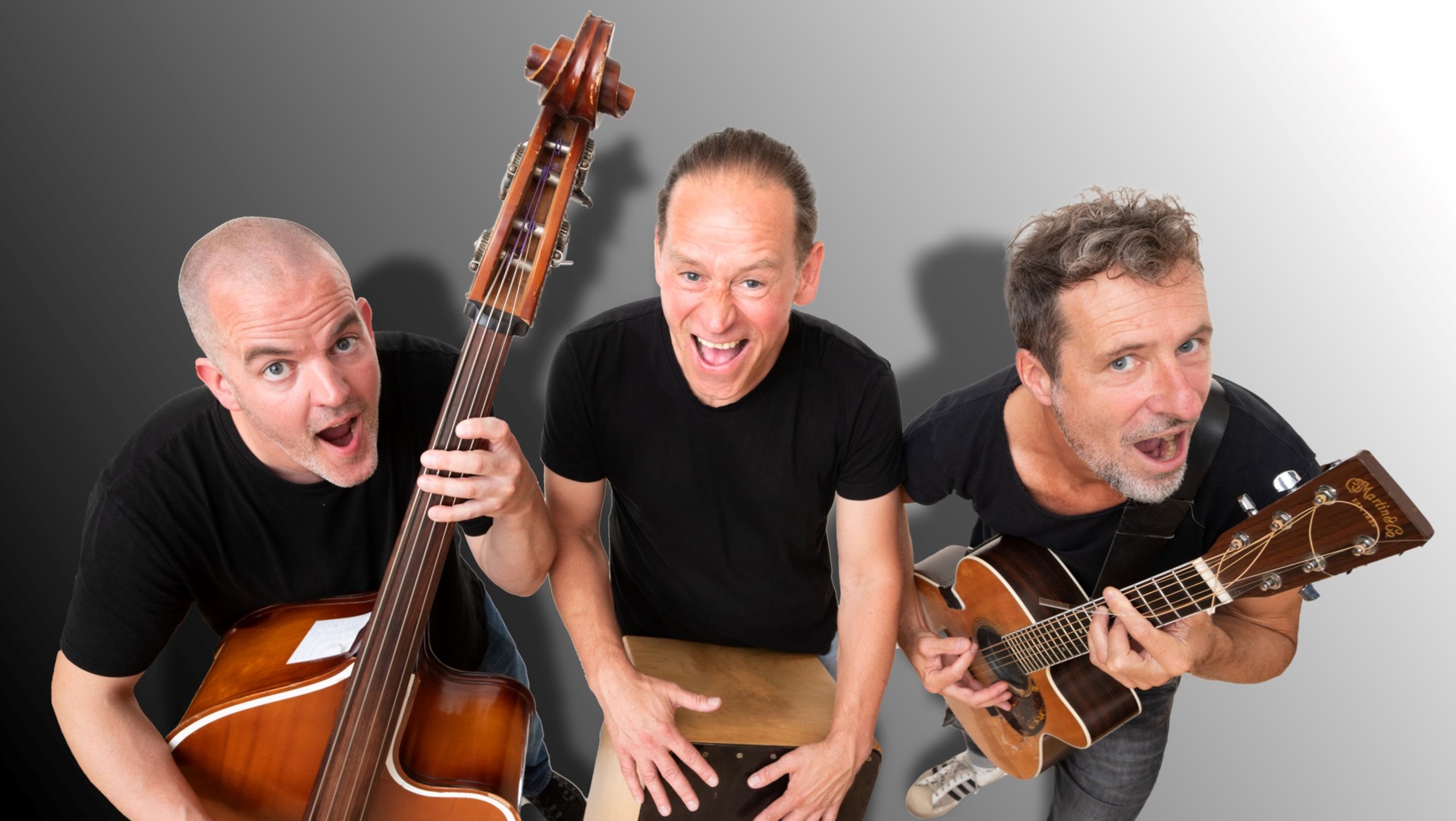 Die drei Bandmitglieder lächeln mit offenem Mund in die Kamera und halten jeweils ihr Instrument in der Hand. Sie sind alle schwarz gekleidet.