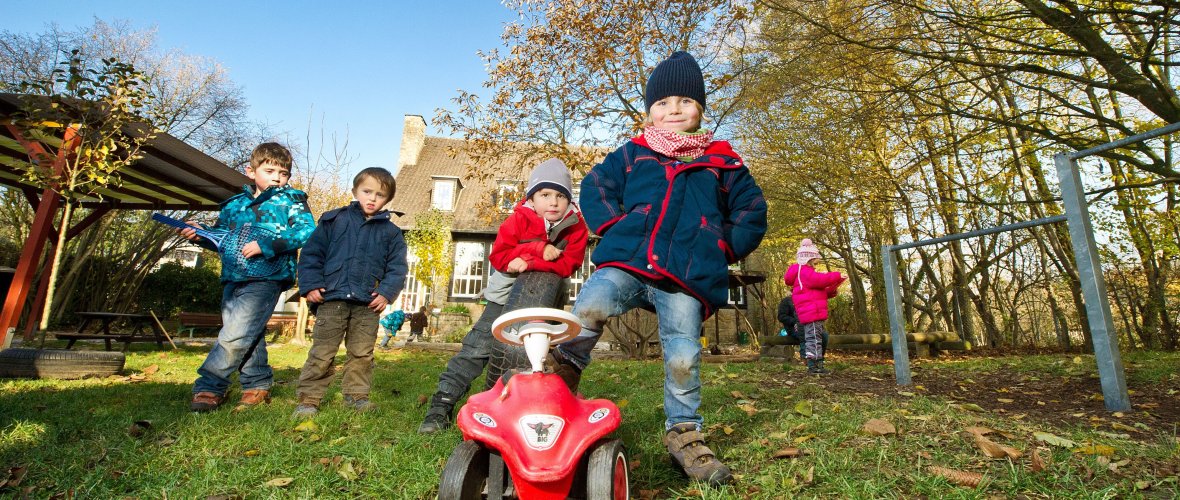 Kinder spielen draußen auf einer Wiese, vorne steht ein Kind mit einem Fuß auf einem roten Bobby Car