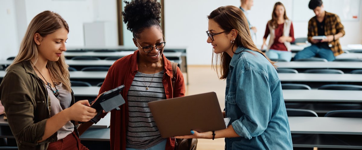 Studentinnen stehen entspannt mit Laptop und Tablet in einem Hörsaal.