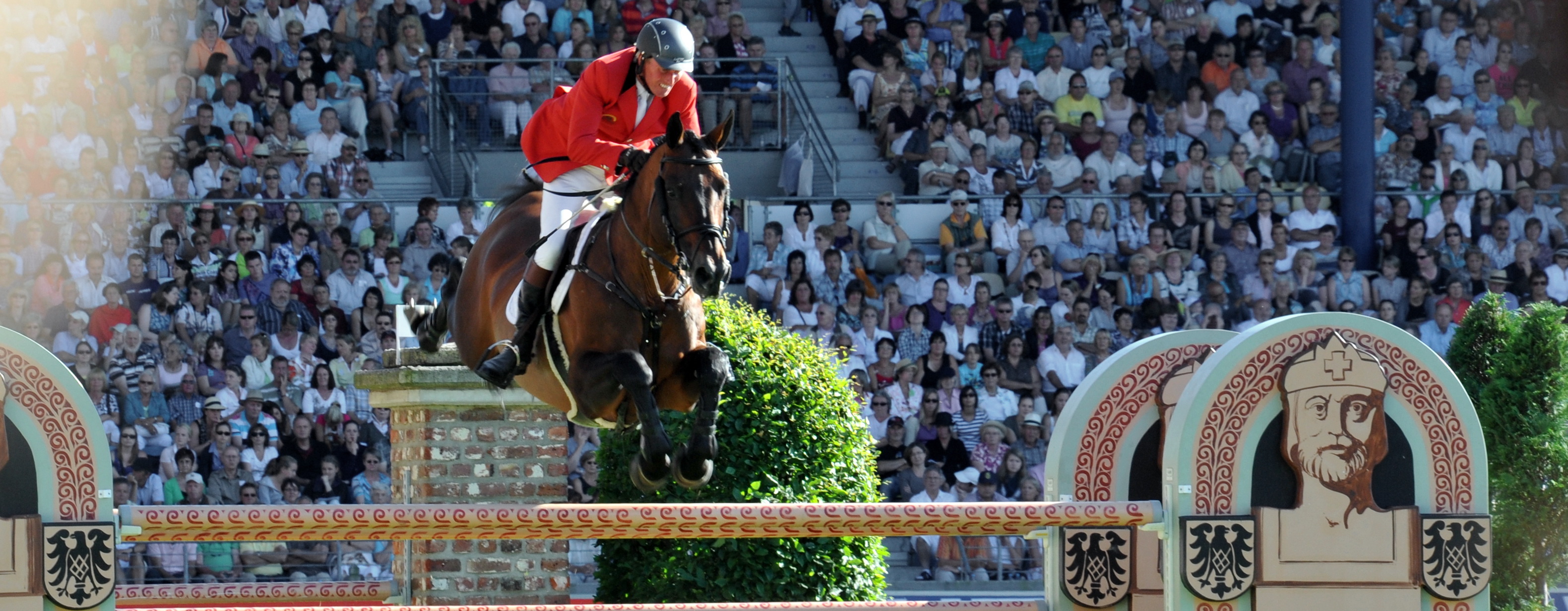 Pferd und Reiter springen über ein Hindernis. Im Hintergrund gut gefüllte Reitstadion in der Soers.