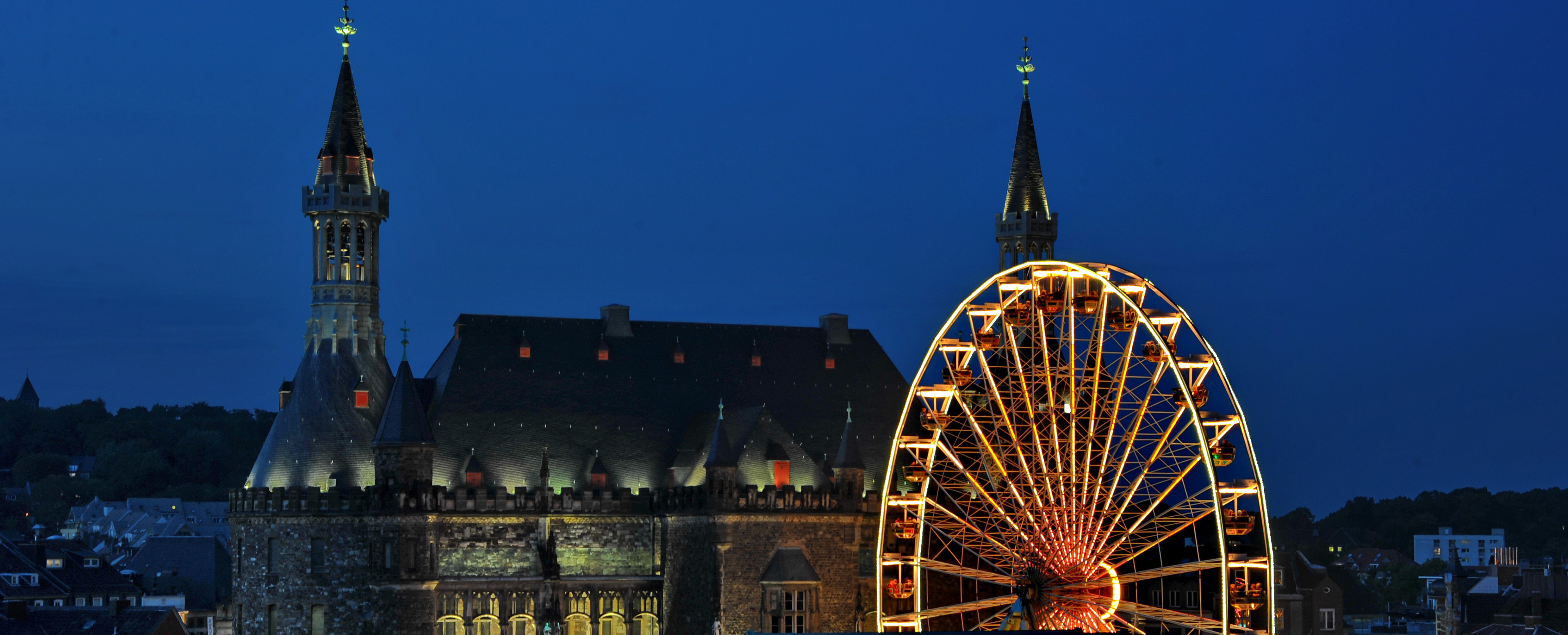 Luftaufnahme: Das Aachener Rathaus bei Nacht, im Vordergrund das hell erleuchtete Riesenrad des Septemberspecials.