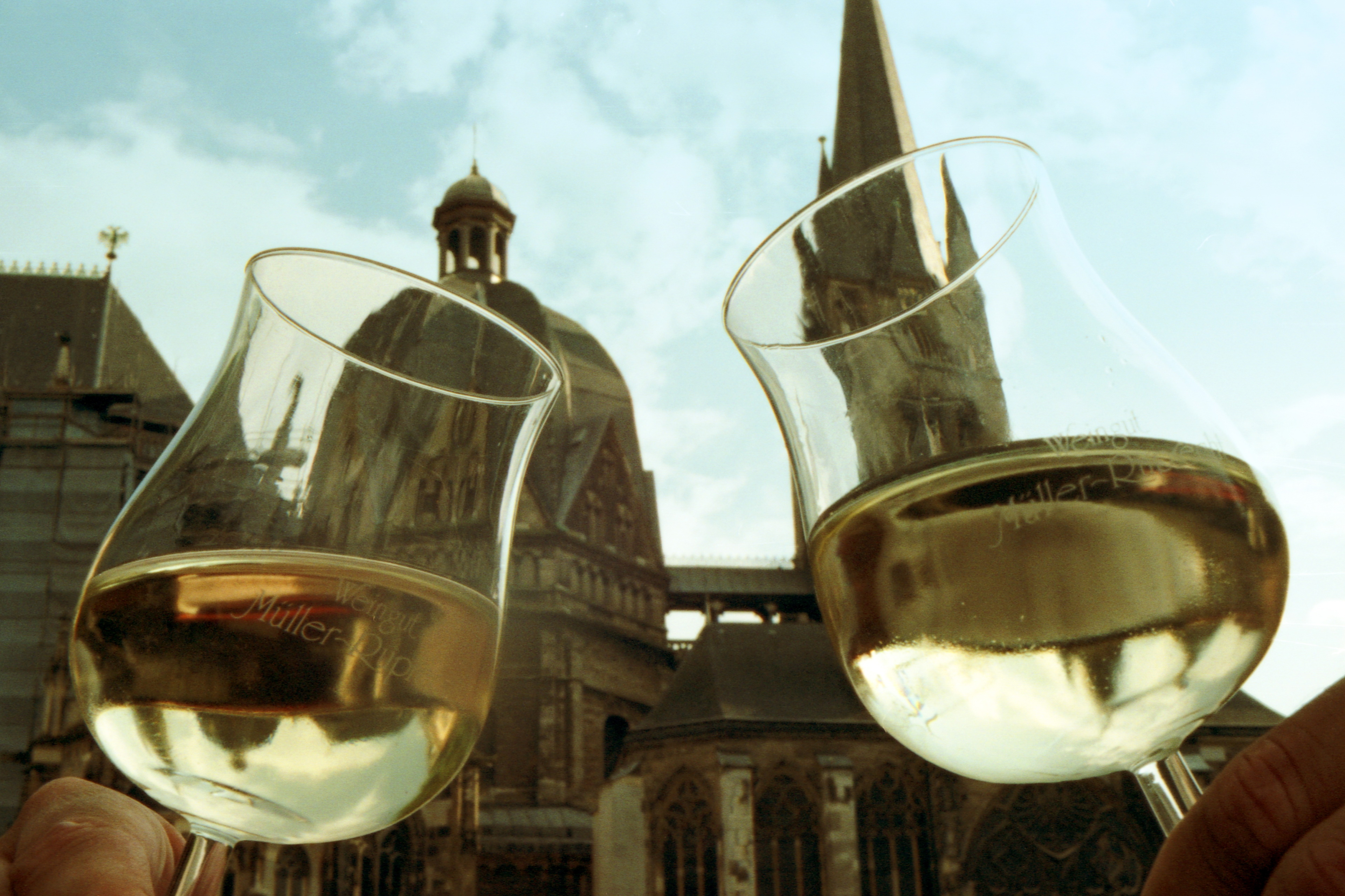 Zwei volle Weingläser werden angestoßen. Im Hintergrund der Aachener Dom.
