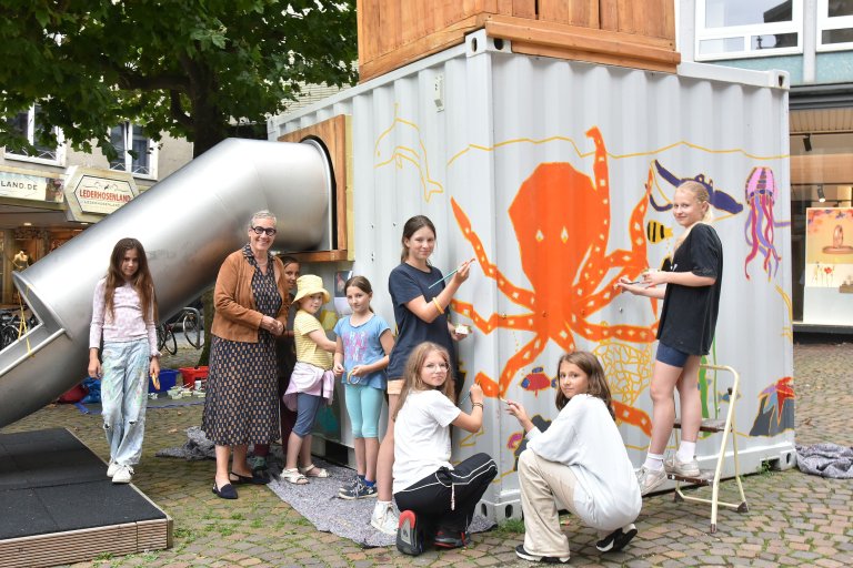 Eine Gruppe von Kindern zusammen mit Oberbürgermeisterin Sibylle Keupen vor einem mit Wassermotiven bemalten Container.