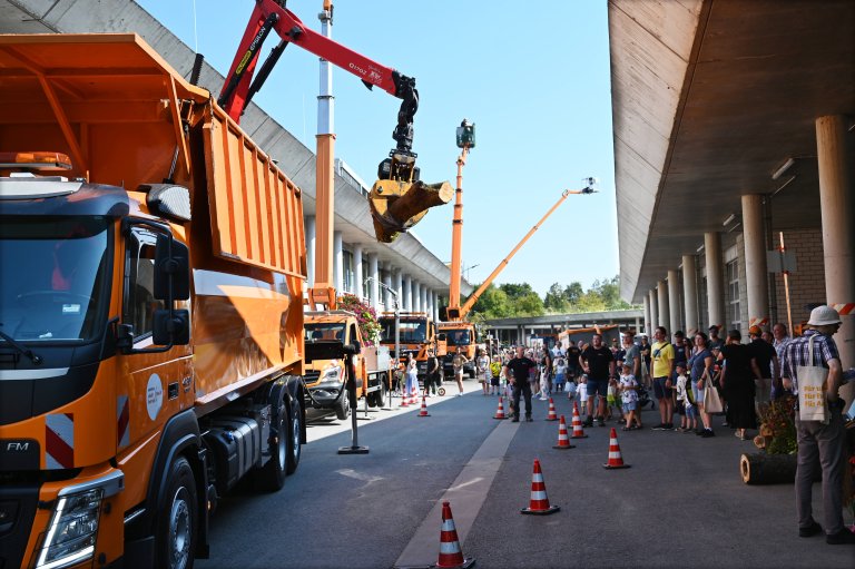 Ein Containerfahrzeug mit einem Kran hebt einen Baumstamm an einer Klaue hoch, während viele Besucher der 25. Jubiläums des Stadtbetriebs zuschauen. Im Hintergrund sind andere Fahrzeuge des Stadtbetriebs zu sehen.