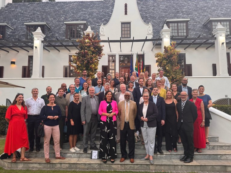 Eine Gruppe Menschen steht auf einer offenen Treppe vor einem großen haus mit Veranda. Im hintergrund die Flaggen von Südafrika, Deutschland und der EU