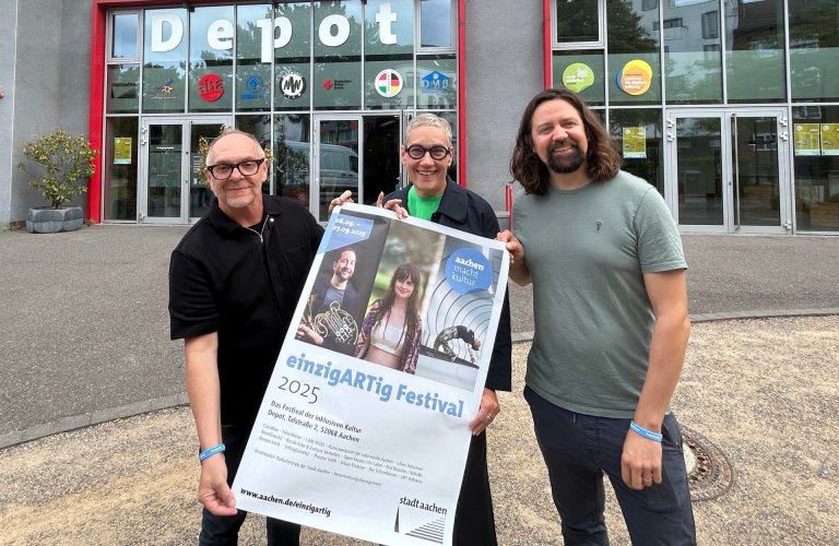 Drei Menschen lachen in die Kamera und halten ein Plakat in den Händen. Im Hintergrund ein Gebäude mit großen Glasfenstern und dem Schriftzug "DEPOT"