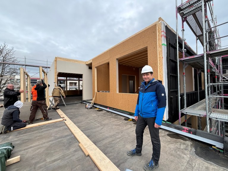 Ein Mann mit Bauhelm steht auf einem Gebäude, welches angebaut wird vor einem entstehendem Gebäudekomplex mit Holzwänden. Im Hintergrund Gerüst und bewölkter Himmel.