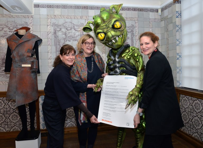 Drei Frauen stehen gemeinsam mit einer grünen Außerirdischen-Figur vor einer gekachelten Wand und halten ein großes Papier in den Händen.